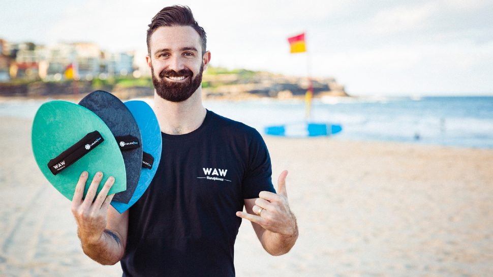 WAW Handplanes Founder Rikki Gibley stands on the beach and proudly holds three handplanes. 