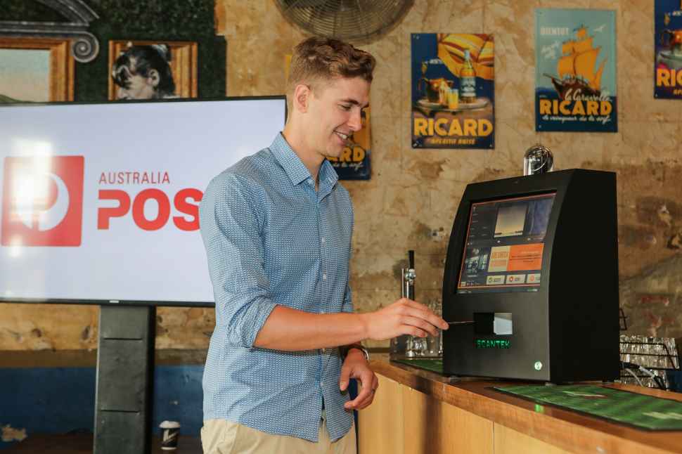 At the launch of event of Digital ID in Melbourne, a young man is scanning his phone in a machine sitting on a bench. Behind him is a screen with the Australia Post logo on it.